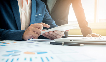 M�os de um homem mexendo no celular, a sua frente est�o um laptop, uma caneta e pap�is com gr�ficos impressos.